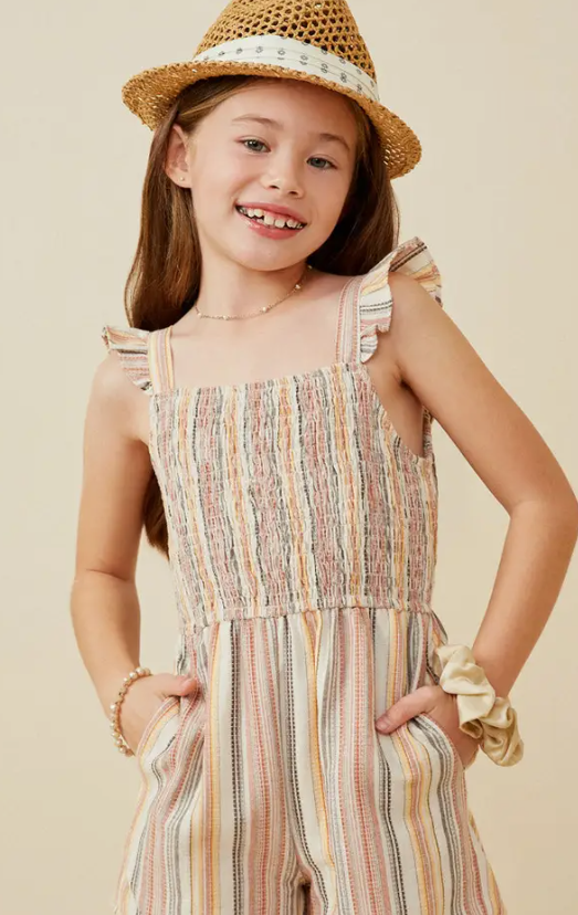 A smiling young girl wears the Hayden LA Tan Ruffle Strap Smocked Romper with her hands in the pockets. She accessorizes with a bracelet and scrunchie, topped with a straw hat, all against a neutral background.