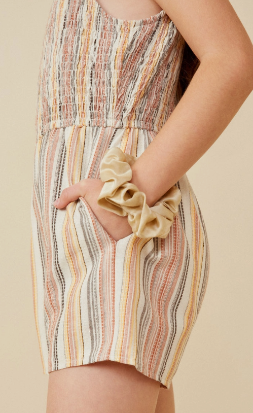 A person wears the Hayden LA Tan Ruffle Strap Smocked Romper, featuring ruffle straps and smocked details, with one hand in their pocket and a beige scrunchie on their wrist.