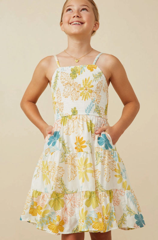 A girl smiles as she stands with her hands in her pockets, wearing the Hayden LA Girls Tropic Smocked Tank Dress— a sleeveless sundress with yellow, green, and blue florals— and looks slightly upward against a neutral background.