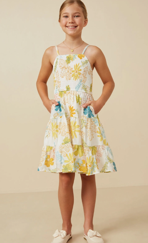 A young girl smiles in the Hayden LA Girls Tropic Smocked Tank Dress, featuring yellow, blue, and green floral patterns. She has her hands in the dress pockets and wears cream bow shoes, posed against a neutral background.