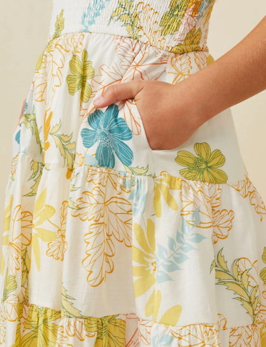 A girl wears the Hayden LA Girls Tropic Smocked Tank Dress, featuring white fabric with colorful floral and leaf patterns, yellow, blue, and green designs, and a gathered ruffled skirt. She stands with one hand in a side pocket.