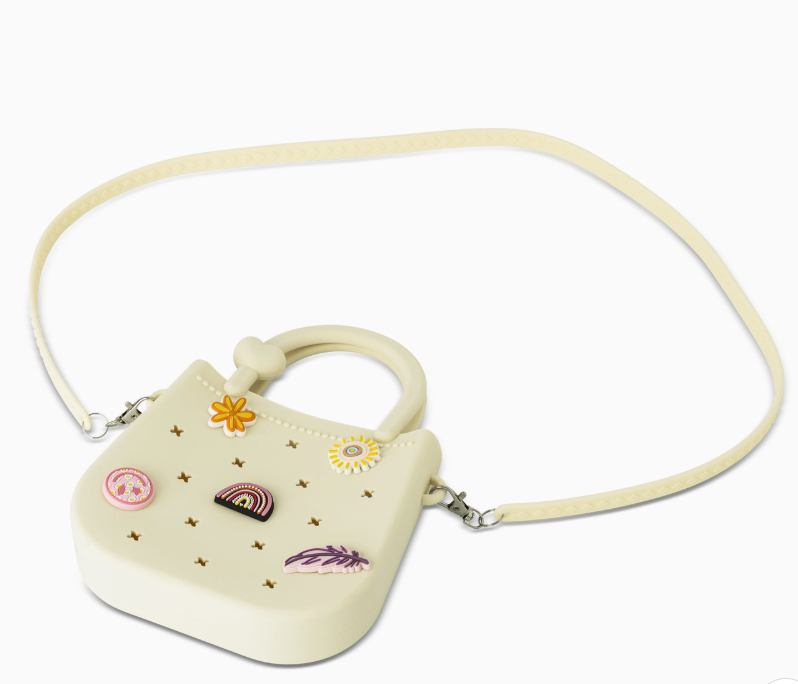 The core home Mini Round Eva Set Purse is a small, cream bag with a long strap, rounded handle, and star-shaped front cutouts. It features colorful pins—rainbow, flowers, and a feather—for playful style.