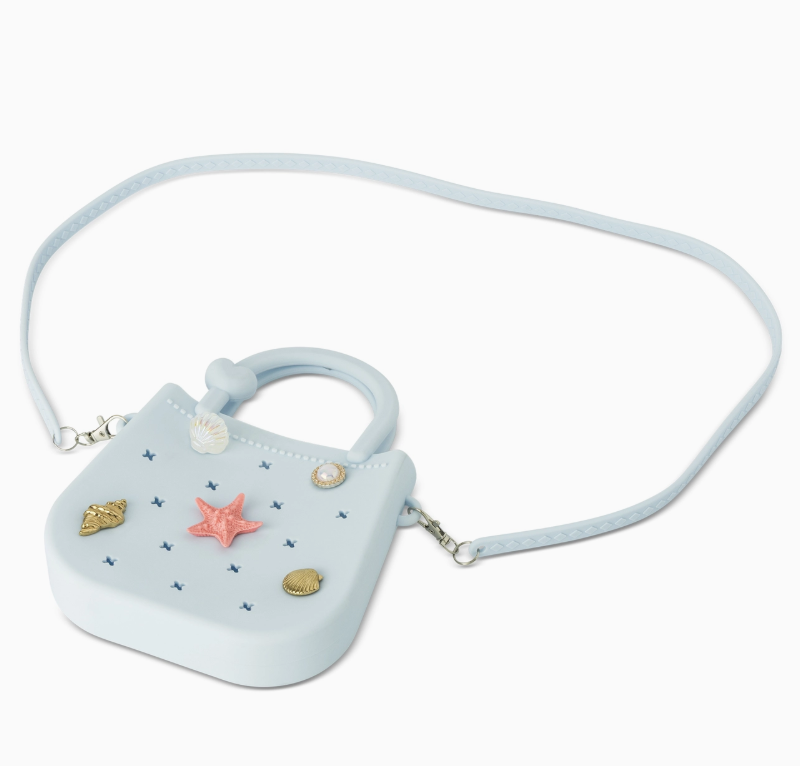 The core home Mini Round Eva Purse - Blue is a small, light blue bag featuring a long strap and decorated with ocean-themed accents like a pink starfish, gold seashells, and small blue crosses.