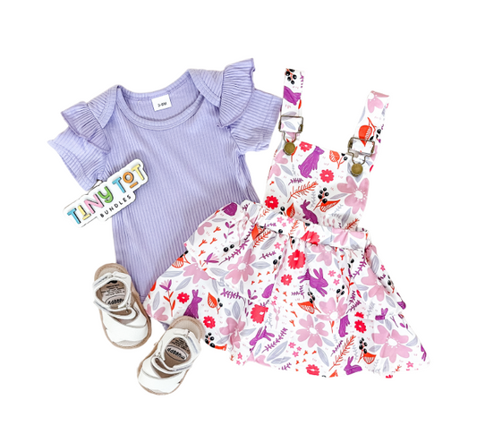 A flat lay of the Tiny Tot Bundles Purple Floral Suspender Set for toddler girls—featuring a lilac ruffled-sleeve top, floral pink and white suspender dress, beige sandals, and a brand sticker on a white background.
