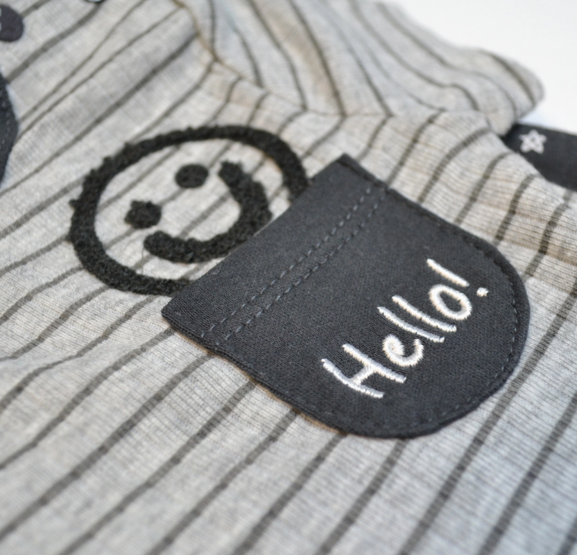 Close-up of Lollipop Dreams’ Hello Happy Bubble Trio: gray and white striped fabric with a small dark pocket embroidered with "Hello!" beside a stitched black smiley face.