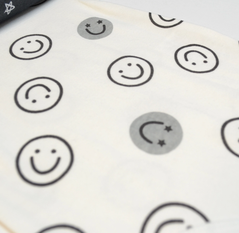 Hello Happy Bubble Trio by Lollipop Dreams is a white fabric featuring a pattern of black smiley faces—some shaded gray with star eyes, others outlined in black with regular eyes.