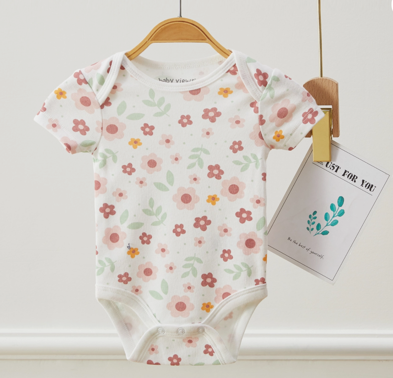 A Lollipop Dreams Flower Power Bubble Trio, a white short-sleeved baby onesie with pink, orange, and green floral patterns, hangs on a wooden hanger beside a card reading JUST FOR YOU with a leafy illustration.