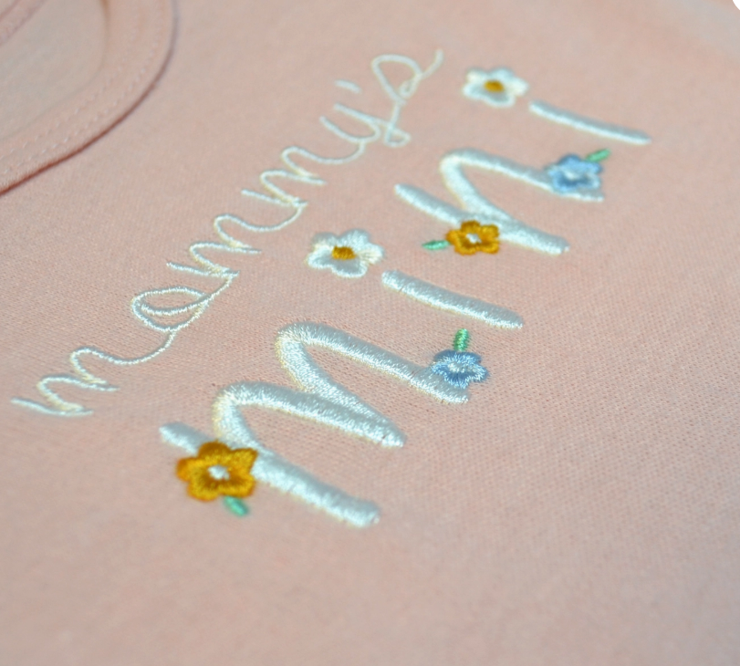 A close-up of pink fabric from the Lollipop Dreams Mommy’s Mini Bubble Trio, featuring "mommys mini" embroidered in white, accented with small yellow and blue flower designs.