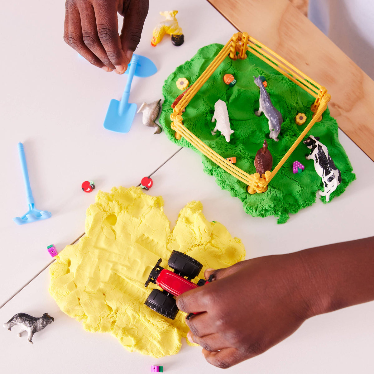 A child’s hands play with the Sensory Pack Farm On the Go Sensory Play Set for Kids by Faber-Castell / Creativity for Kids, featuring a toy tractor, blue shovel, yellow and green playdough, toy farm animals, a fenced pen, and tiny apples.
