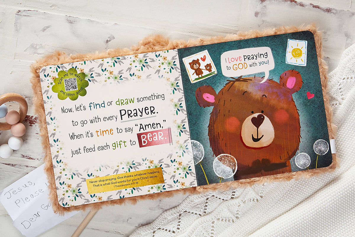 An open Prayer Bear Book by BroadStreet Publishing Group, LLC shows a bear illustration and colorful text inviting children to draw something to accompany each prayer. Next to the book, on a white textured cloth, lies a handwritten note that reads, “Jesus, please Dear &...”.