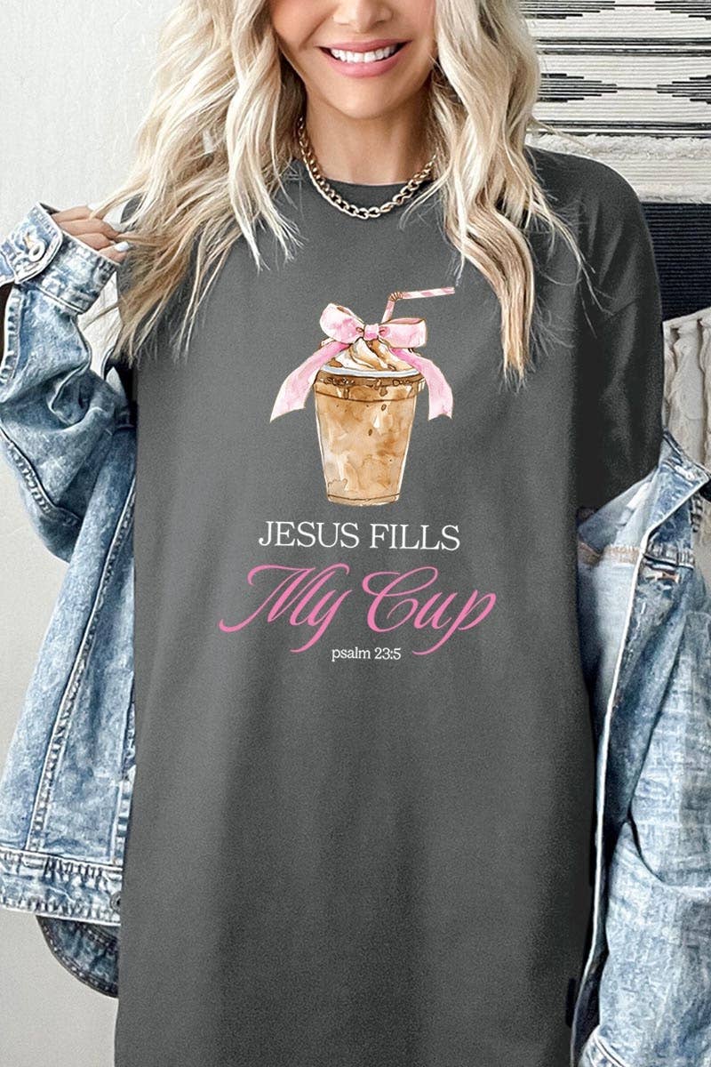 A blonde woman wears a COLORBEAR Jesus Fills My Cup Graphic Cotton Tee, featuring an iced coffee illustration, “JESUS FILLS My Cup” in white and pink lettering, and Psalm 23:5 below. She has a denim jacket draped over her shoulders.