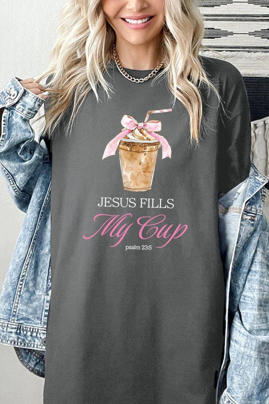A blonde woman wears a COLORBEAR Jesus Fills My Cup Graphic Cotton Tee, featuring an iced coffee illustration, “JESUS FILLS My Cup” in white and pink lettering, and Psalm 23:5 below. She has a denim jacket draped over her shoulders.