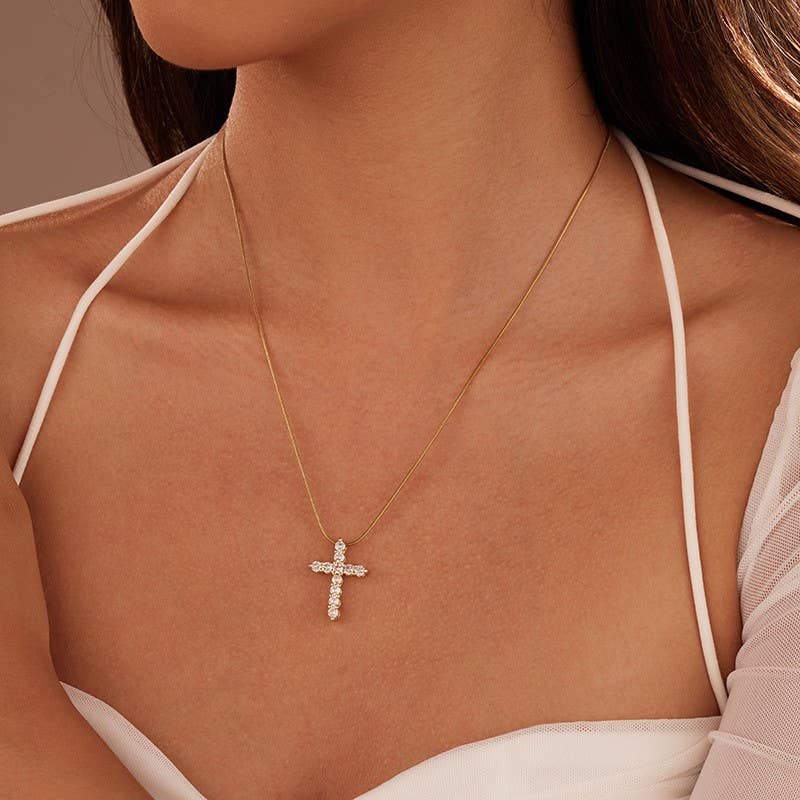 A woman wearing a white spaghetti strap top and the JOSSLYN by wall to wall Tarnish Free Cross Necklace, featuring a gold chain and sparkling cross pendant, shown from shoulders to chin.
