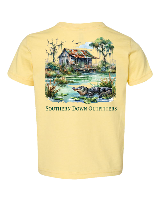 The Swamp Shack Toddler Tee by Southern Down Outfitters is a light yellow t-shirt with an alligator illustration by a pond, old wooden cabin, trees, and birds. Features "SOUTHERN DOWN OUTFITTERS" text.