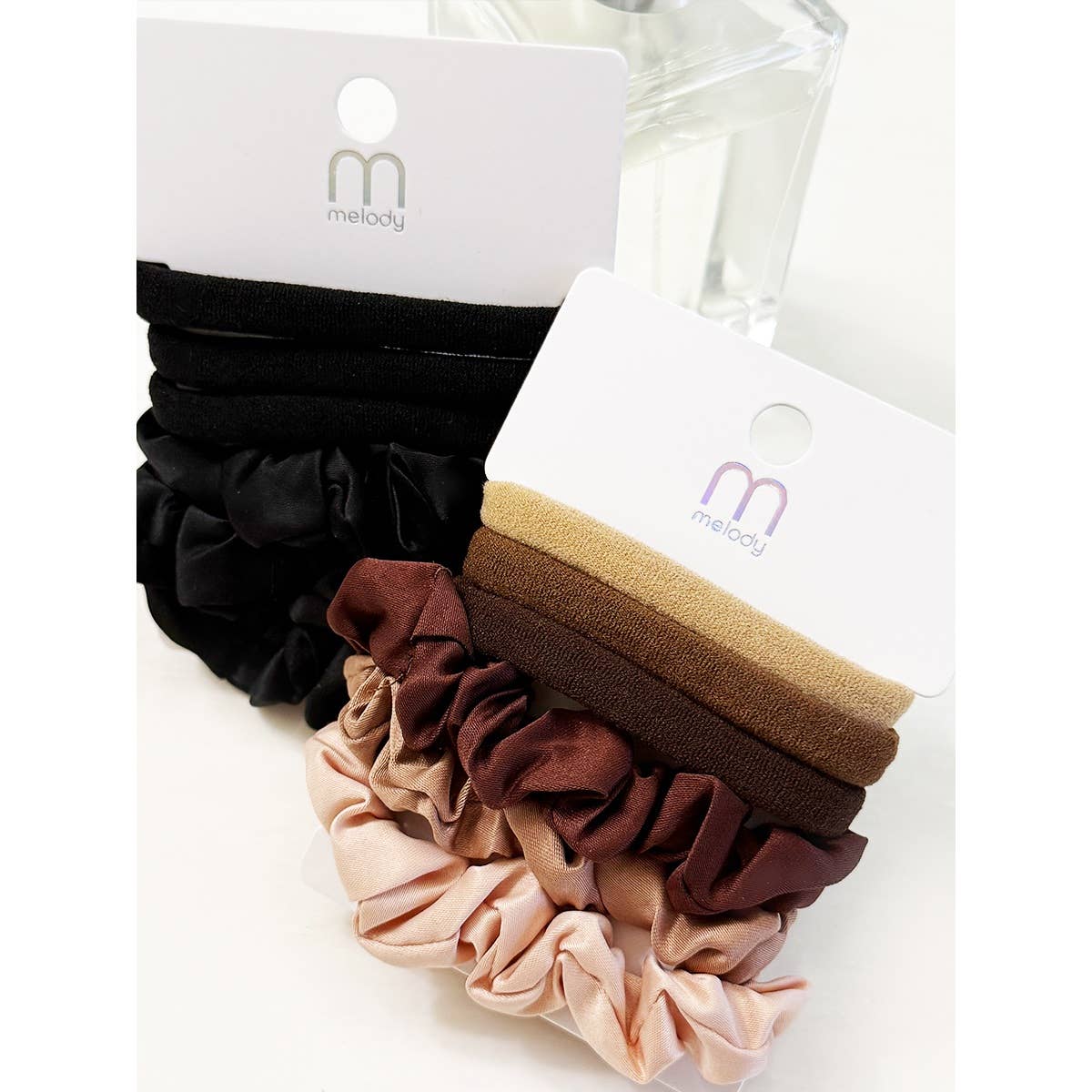 Two sets from the Love and Repeat Assorted Hair Tie & Scrunchie Set, featuring black, brown, tan, and beige shades, are displayed on white cards marked with “melody,” with a blurred perfume bottle in the background.