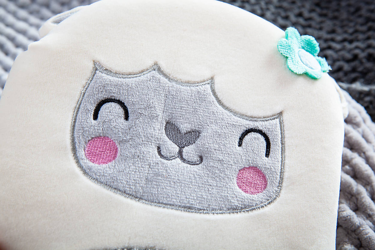 A close-up of the Squish and Snugg Lamb Book by BroadStreet Publishing Group, LLC, highlighting the lamb’s smiling face with embroidered eyes, rosy cheeks, and a blue flower on its head. The background features a textured gray fabric.