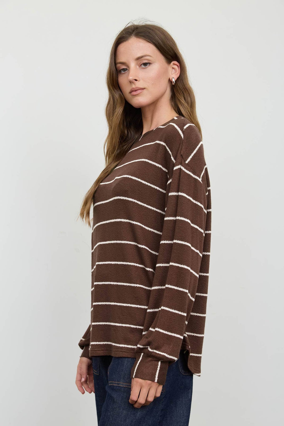 A woman with long brown hair wears the Blu Pepper Brown Stripe Cuffed Knit Top paired with blue jeans, standing sideways and looking at the camera against a plain light background.