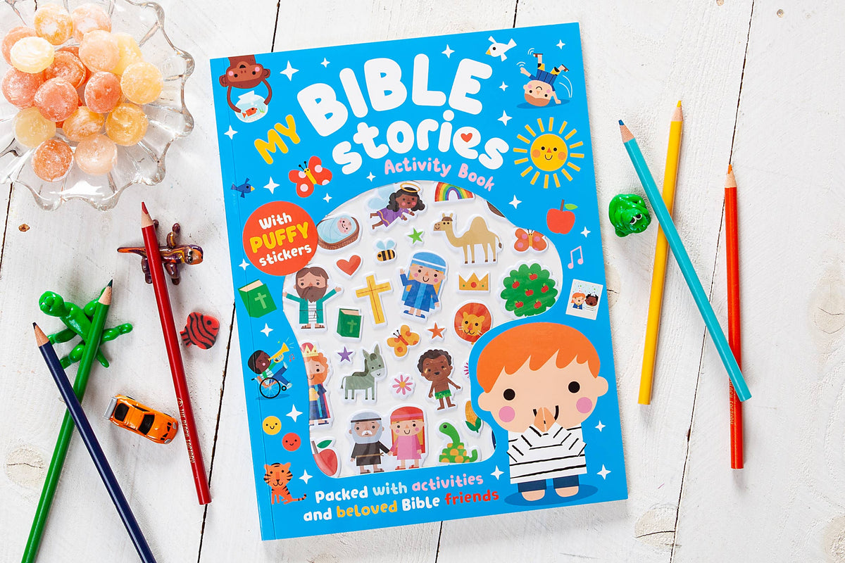 The My Bible Stories Activity Book by BroadStreet Publishing Group, LLC features colorful illustrations, puffy stickers, pencils, and small toys displayed on a white wooden surface. Surrounded by candies, the activity book promises engaging activities and introduces children to Bible friends.