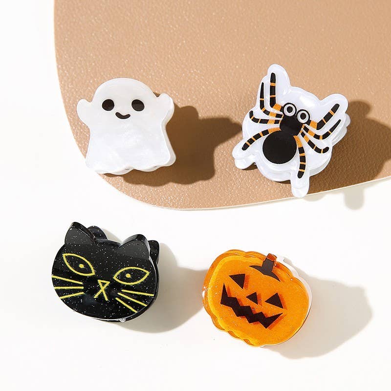 Four Halloween-themed mini hair clips from JOSSLYN by wall to wall—a white ghost, a spider with striped legs, a black cat with yellow eyes, and an orange jack-o-lantern—arranged on a tan and white surface.