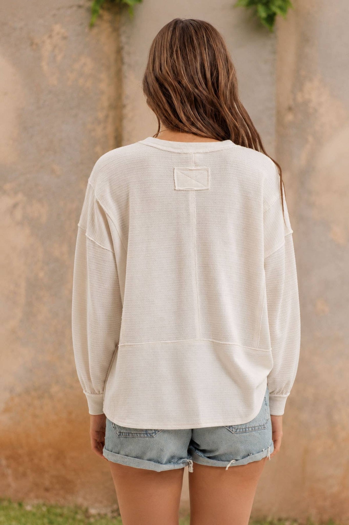 A person with long brown hair stands outdoors facing away, wearing the Blu Pepper Ivory Knit Long Sleeve Top and light blue denim shorts. The background features a textured beige wall with greenery at the top.