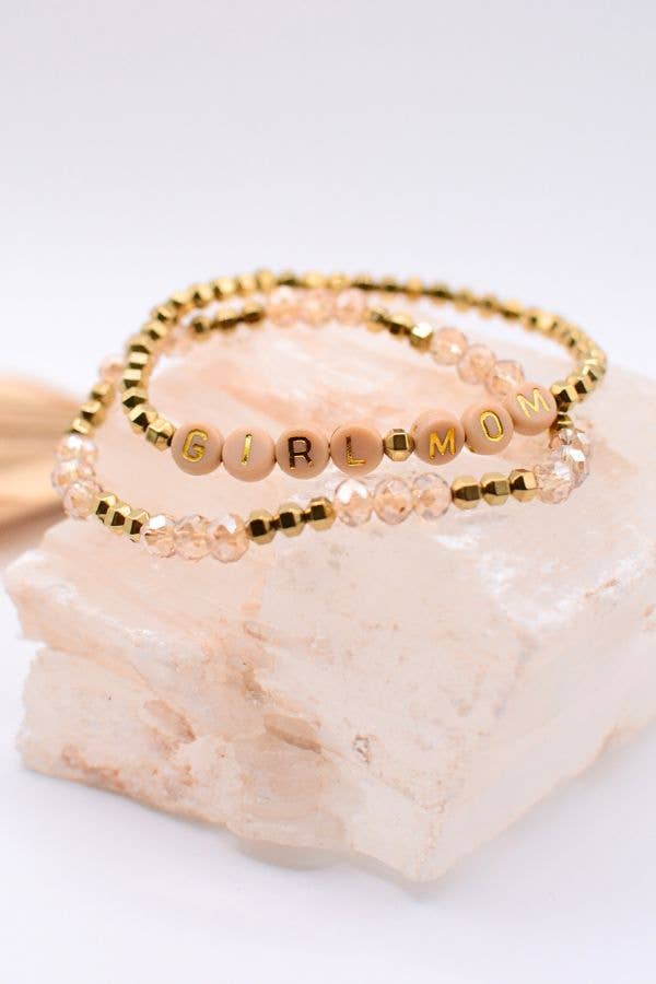 The Girl Mom Bracelet Set by Two and Crew - Faith Based Jewelry & Gifts features two stacked beaded bracelets: one with gold beads, and the other with pink and clear beads accented by letter beads spelling “GIRL MOM.” Both bracelets are displayed on a light, textured stone surface.