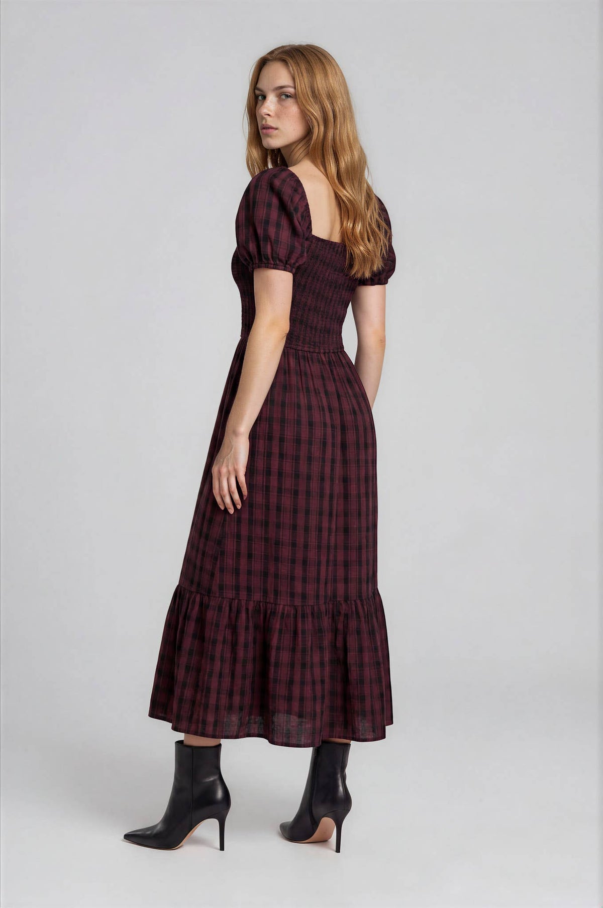 A person wearing a Blu Pepper Burgundy Plaid Midi Dress featuring a smocked bodice, puff sleeves, and a tiered skirt, paired with black heeled boots.