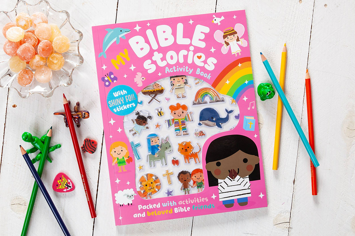 A vibrant My Bible Stories Activity Book from BroadStreet Publishing Group, LLC sits on a white table, surrounded by colored pencils, candies, and small animal toys. The cover showcases cartoon Bible characters and a rainbow.