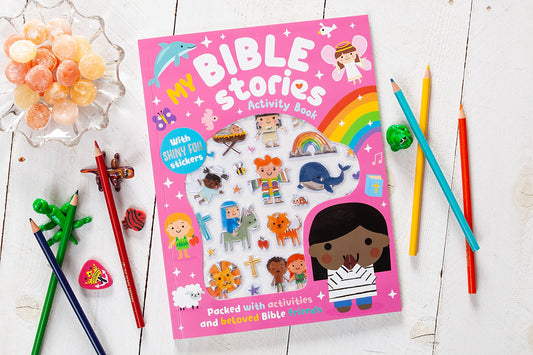 A vibrant My Bible Stories Activity Book from BroadStreet Publishing Group, LLC sits on a white table, surrounded by colored pencils, candies, and small animal toys. The cover showcases cartoon Bible characters and a rainbow.
