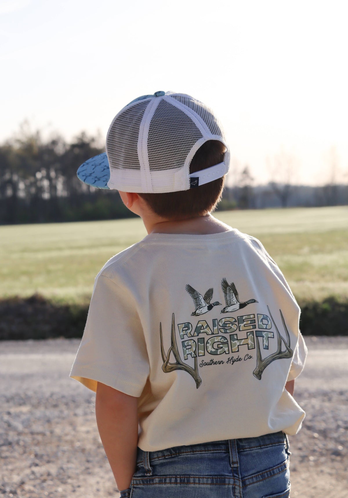 A young boy wears a Raised Right Tee, featuring a graphic of two ducks and antlers above the text Raised Right, and a backwards trucker hat.