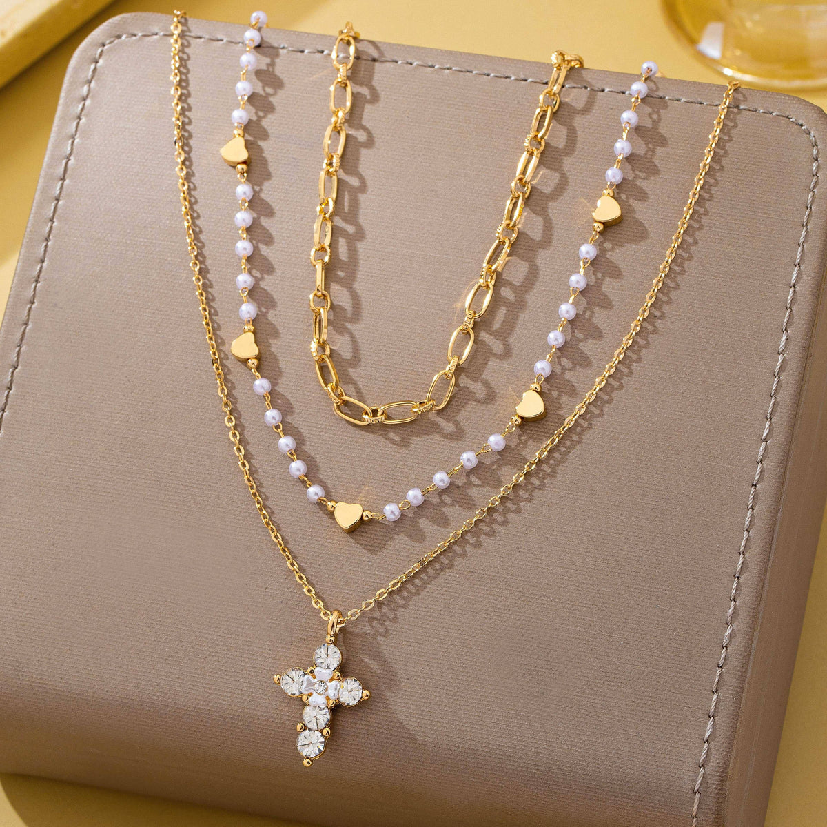 The JOSSLYN by wall to wall Multi-Strand Pearl & Crystal Cross Necklace is displayed on a taupe jewelry box, featuring three layered strands: one adorned with a crystal cross pendant, one with pearl-like beads and gold hearts, and another with chunky gold links.