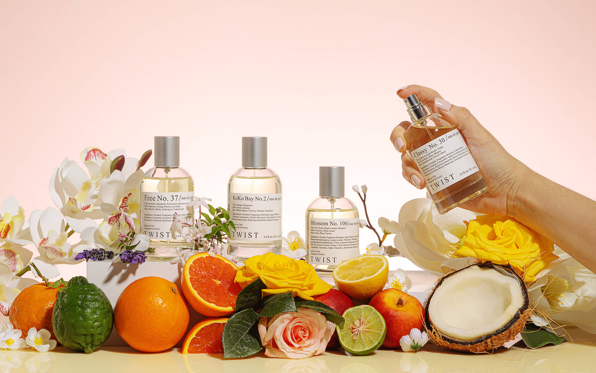 Four bottles of House of Twist Sun Kissed No. 115 Perfume are artfully arranged among fresh flowers, oranges, limes, and a halved coconut—capturing the vibrant essence of summer. A hand is shown misting one bottle into the air, hinting at the allure and long-lasting fragrance designed for women. The backdrop features a delicate gradient of pale pink and orange hues.
