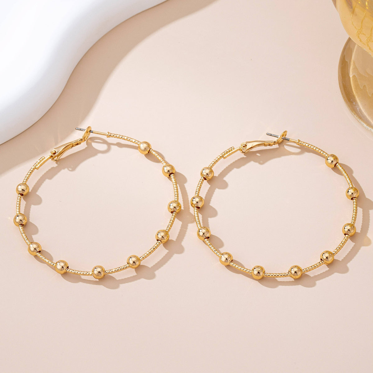 A pair of JOSSLYN by wall to wall Gold Ball Beaded Hoop Earrings, featuring large gold hoops adorned with evenly spaced gold beads, displayed on a beige surface with soft shadows and hints of a white and yellow decorative object in the corners.