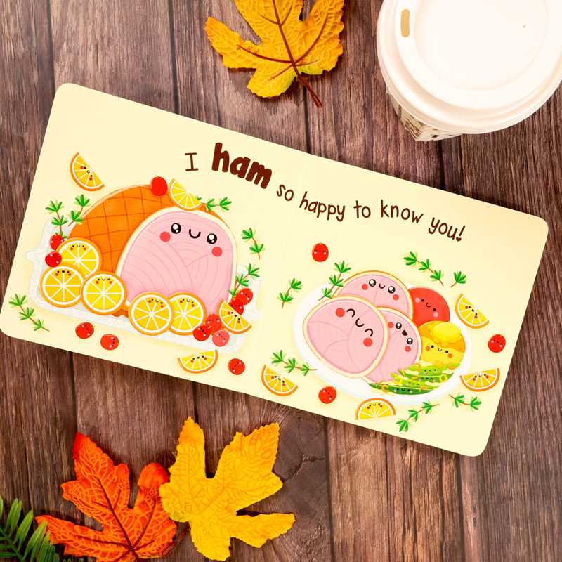 The Pumpkin Spice & Everything Nice card by Dienesa Le from Simon & Schuster features a charming design of smiling ham slices and fruits, with the text “I ham so happy to know you!” The card is surrounded by fall leaves and a cup with a white lid, all placed on a wooden surface.