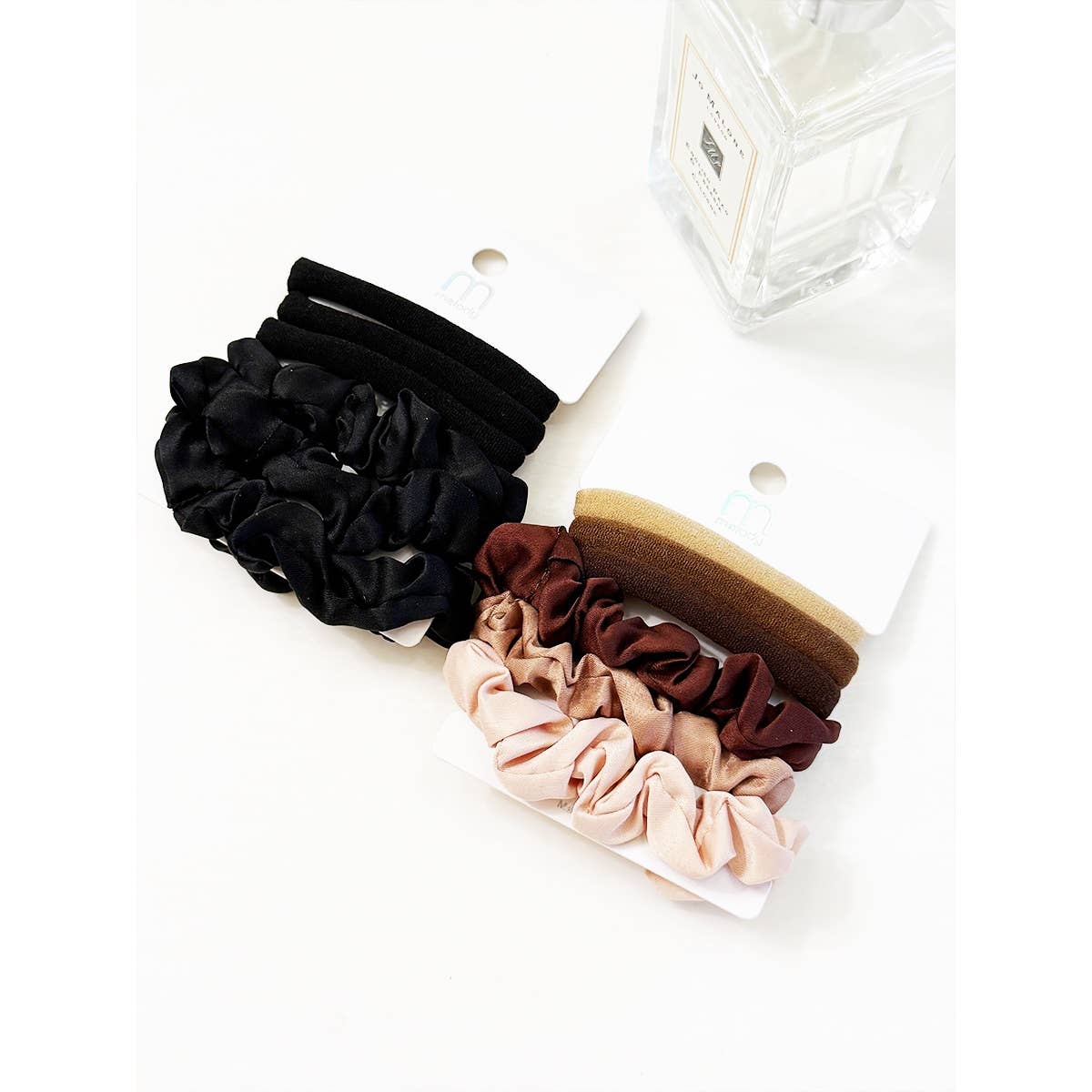 Two Love and Repeat Assorted Hair Tie & Scrunchie Sets in black, brown, and beige tones are displayed on white cards beside a clear glass perfume bottle on a white surface.