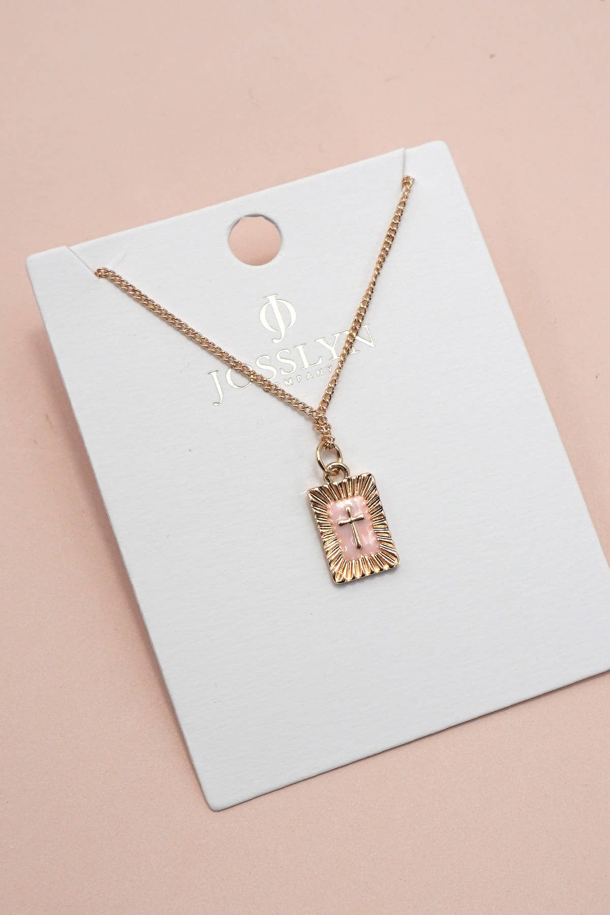 A Sunburst Square Cross Pendant Necklace by JOSSLYN by wall to wall, featuring a gold chain and a rectangular pendant with a pink cross, displayed on a white Josslyn brand card against a light pink background.