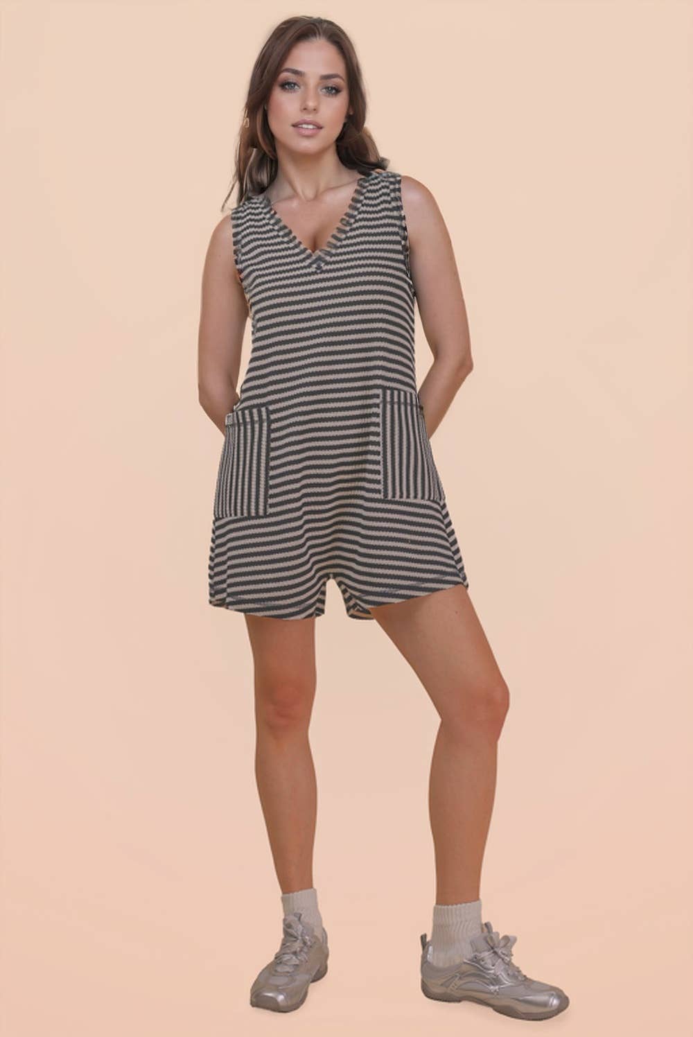 A woman stands against a plain beige background, wearing the VERY J Striped Knit Casual Romper with front pockets, paired with gray socks and metallic silver shoes. She has long brown hair and a relaxed, confident expression.