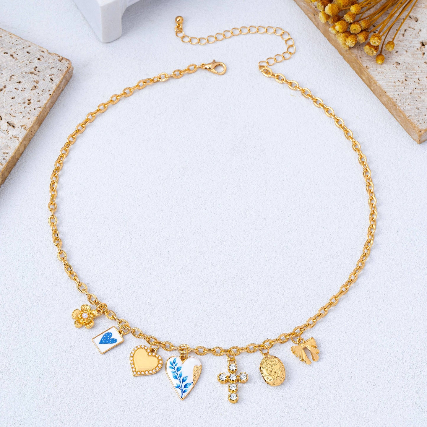 The Blue Vintage Coquette Charm Necklace by JOSSLYN by wall to wall features seven gold charms—a flower, butterfly, heart, leaf, crystal cross, oval medallion, and hand—elegantly displayed on a textured surface with floral accents nearby.