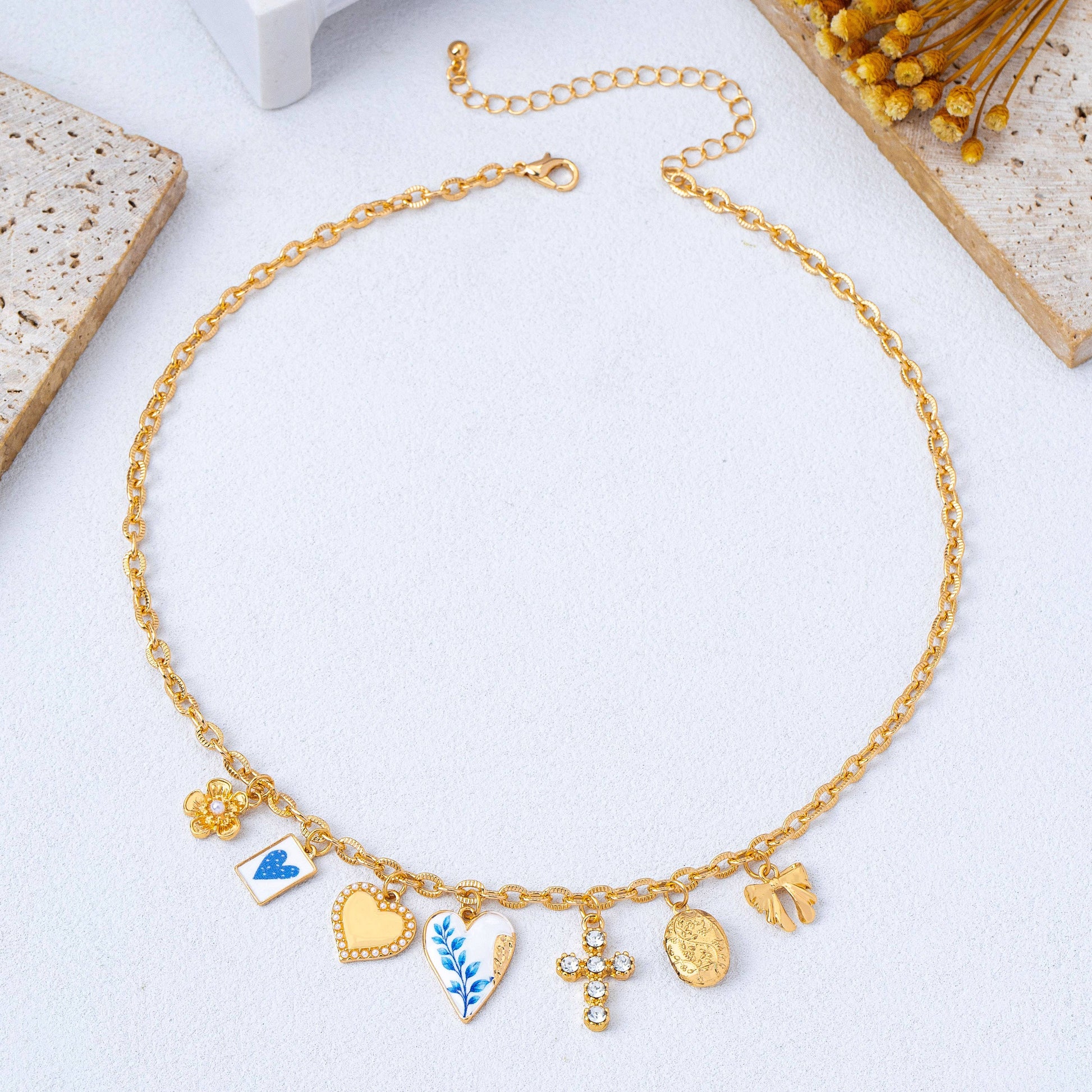 The Blue Vintage Coquette Charm Necklace by JOSSLYN by wall to wall features seven gold charms—a flower, butterfly, heart, leaf, crystal cross, oval medallion, and hand—elegantly displayed on a textured surface with floral accents nearby.