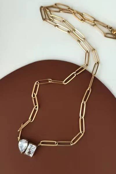 Gold Rhin Necklace by JOSSLYN by wall to wall, a striking gold chain with large elongated links and two clear gemstone pendants, displayed on a brown and cream background.