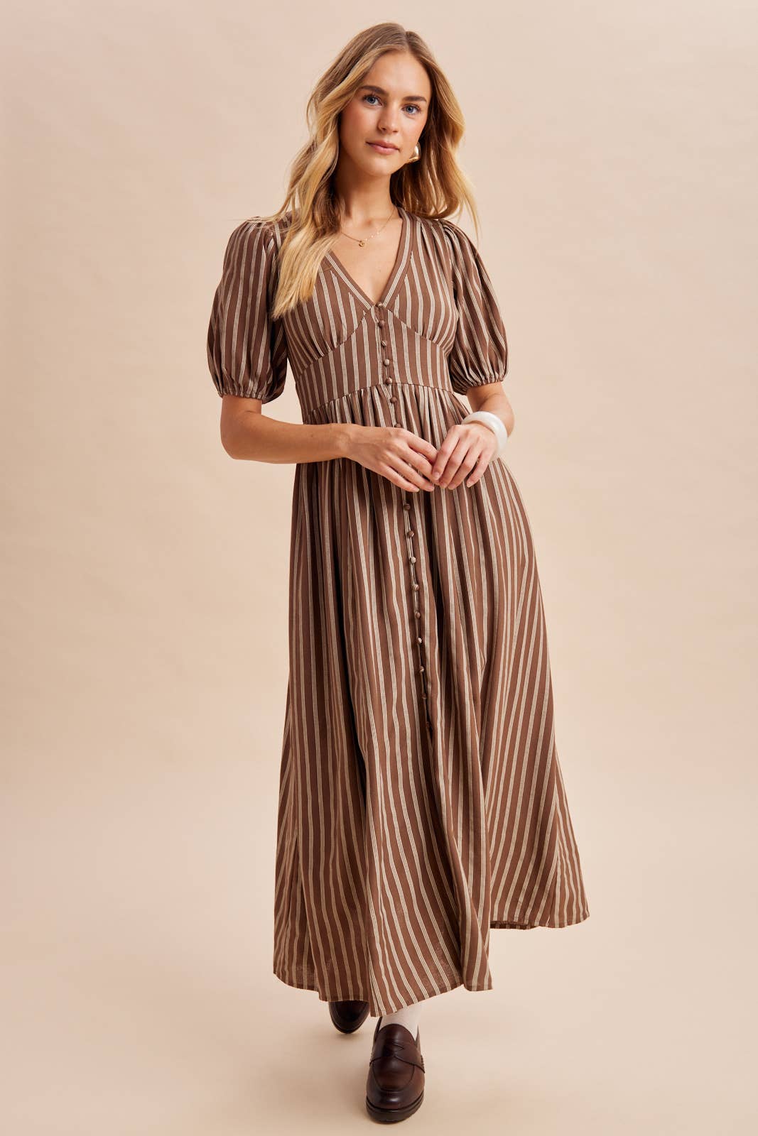A woman wears an In Loom Stripe Button Down Maxi Dress in a brown and white striped pattern with a V-neck, puff sleeves, and front button closure.