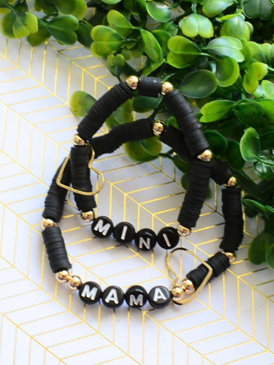 The Mama Mini Bracelet Set - Black by Two and Crew - Faith Based Jewelry & Gifts features two black beaded bracelets with gold accents and heart charms—one spells MINI, the other MAMA—beautifully displayed on a white and gold patterned surface with green leaves in the background.