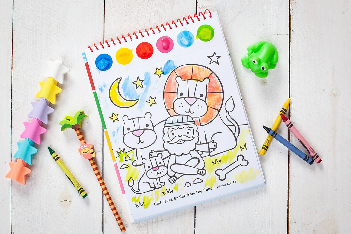 A partially colored page from the Cozy Club Bible Friends Art Pad Book by BroadStreet Publishing Group, LLC features Daniel with lions, a moon, and stars. Nearby on the white wooden table are crayons, a frog toy, and colorful star-shaped items.