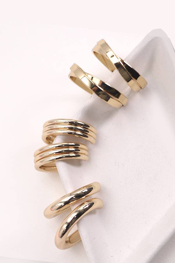 Five gold earrings from the JOSSLYN by wall to wall Multi Hoop Trio Earring Set are arranged on the corner of a white, angular surface against a pale background. The earrings feature smooth, shiny finishes and modern, minimalist designs.