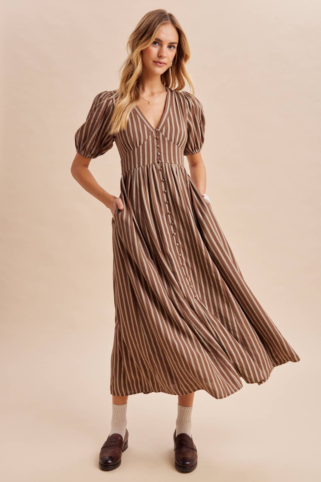 A woman wears an In Loom Stripe Button Down Maxi Dress in brown and beige with vertical stripes, puff sleeves, a v-neckline, and a button-front closure, paired with dark brown loafers.