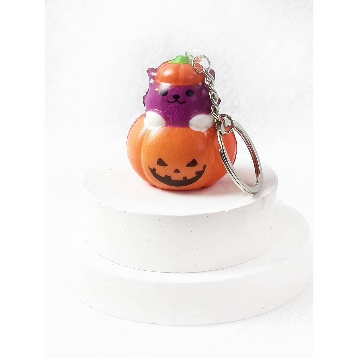 A Love and Repeat Halloween Squishy Keychain featuring a small, squishy purple cat character sitting inside an orange jack-o-lantern, attached to a metal keychain.