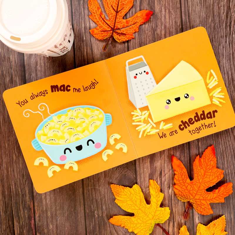 An open copy of “Pumpkin Spice & Everything Nice” by Dienesa Le from Simon & Schuster displays adorable cartoon macaroni, cheese, and a grater with smiley faces. The pages read, “You always mac me laugh! We are cheddar together!” Autumn leaves and a drink rest nearby on a wooden surface.