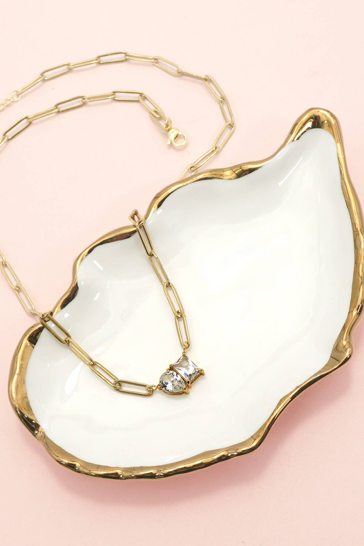 The Gold Rhin Necklace by JOSSLYN by wall to wall, featuring a pendant adorned with two clear stones, rests on a white, gold-rimmed, shell-shaped dish against a light pink background.