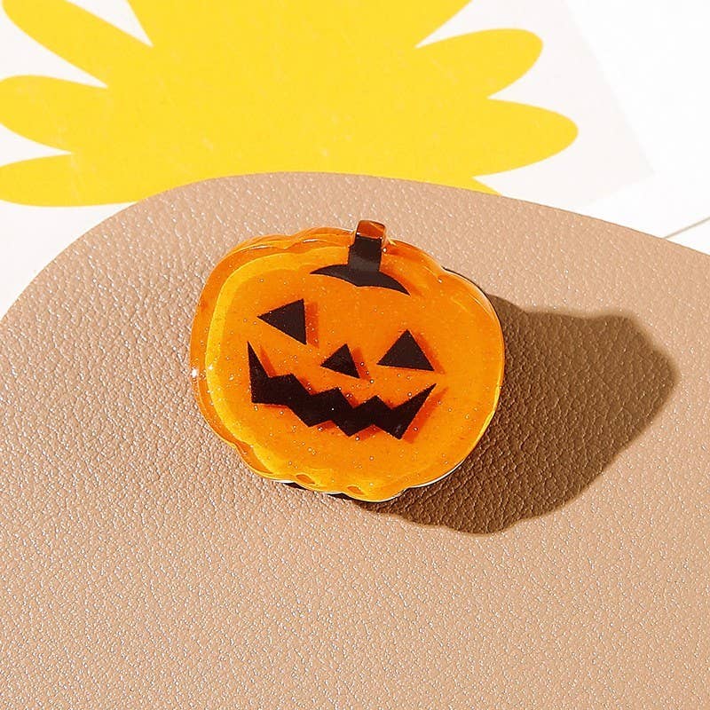 A cute orange Jack-o-lantern hair clip from the Halloween Ghost Theme Mini Hair Clips 4-Pack by JOSSLYN by wall to wall rests on a textured beige surface, casting a shadow.