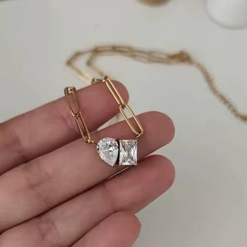 A hand holds the JOSSLYN by wall to wall Gold Rhin Necklace, featuring two large clear gemstone pendants—one pear-shaped and one rectangular—against a blurred white background.