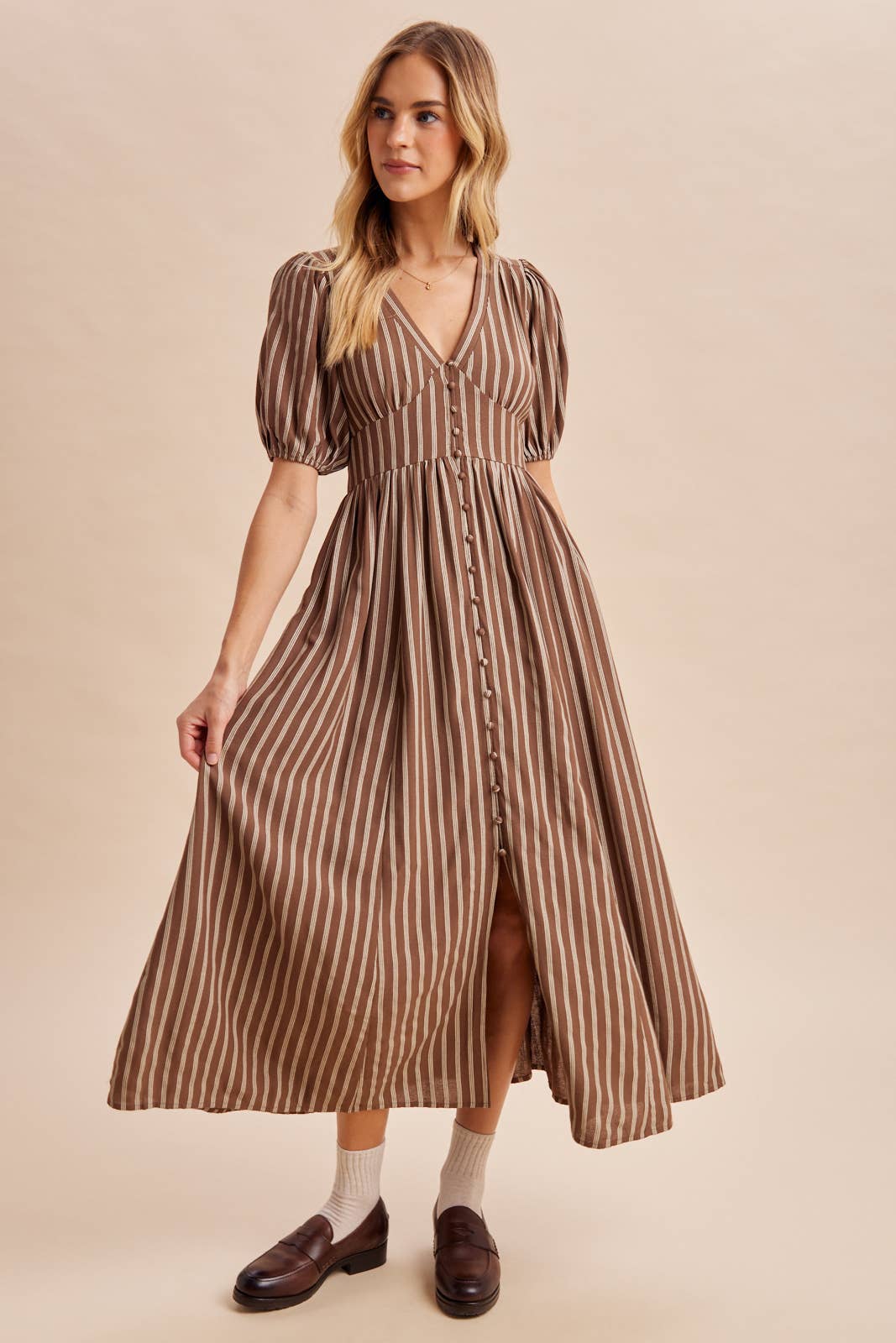 A woman wears an In Loom Stripe Button Down Maxi Dress, a brown and white striped V-neck style with puff sleeves and a front button placket.